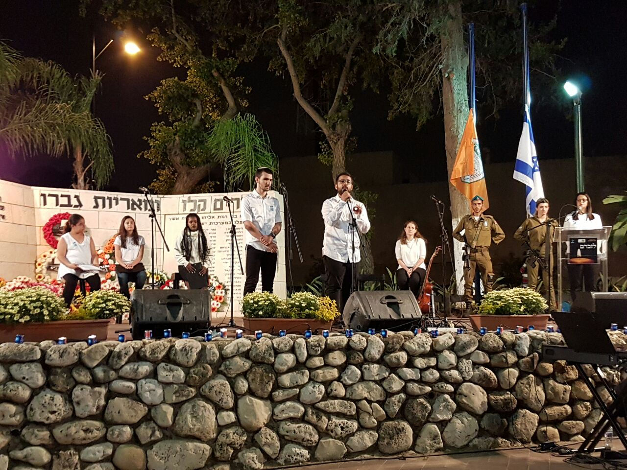טקס יום הזיכרון שני בחורים צעירי עומדים ומדברים לרמקול ומאחוריהם נשים ובחורים צעירים עומדים